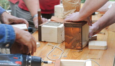 Auf einem Holztisch sieht man Hände von unterschiedlichen Menschen, welche Wildbienen-Nisthilfen aus Holz und Schilfrohr bauen.
