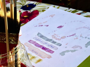 Auf einem weißen Blatt-Papier wurden verschieden Gauche Farben aus Pflanzenfarben gemalt, links im Bild ein Glas Wasser mit Pinseln